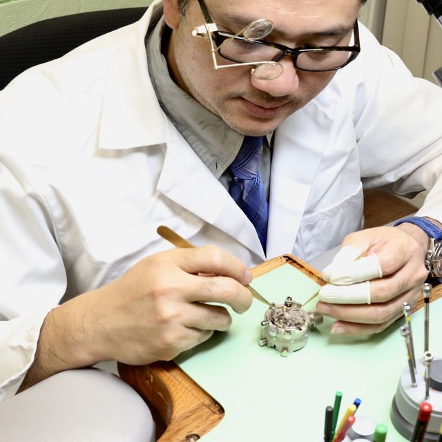 Watch Zone's watchmaker in a white coat, working on a watch movement with tweezers and finger cots beside a bench microscope