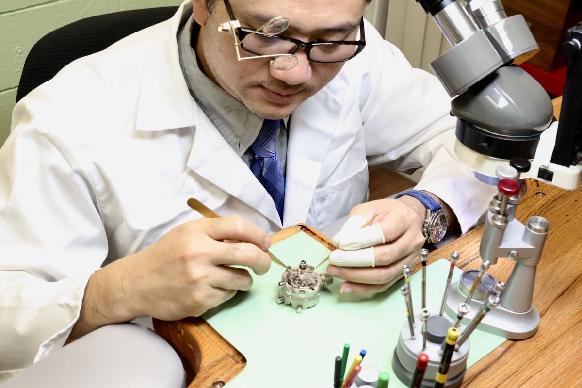 The watchmaker working on a movement held in a movement holder, beside a microscope and a stand of screwdrivers