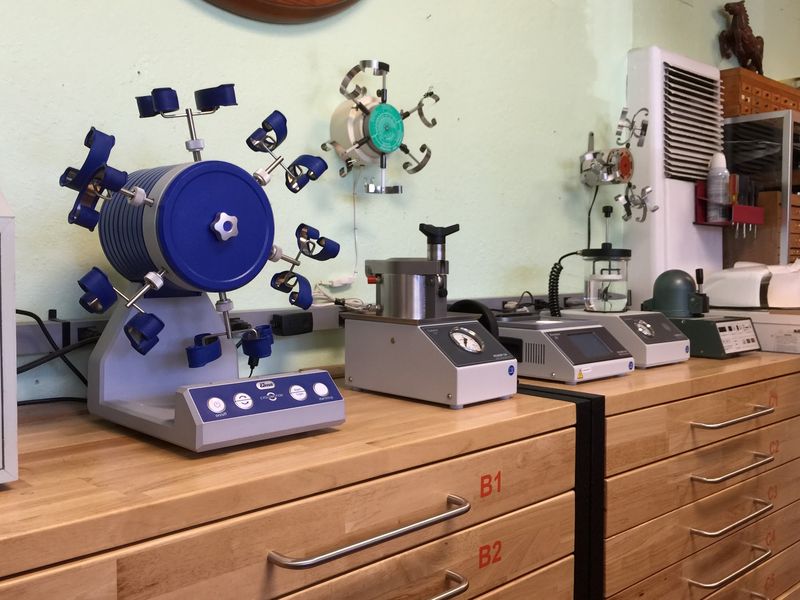 A blue Elma watch cleaning machine and a row of water-resistance testers on wooden workbench drawers