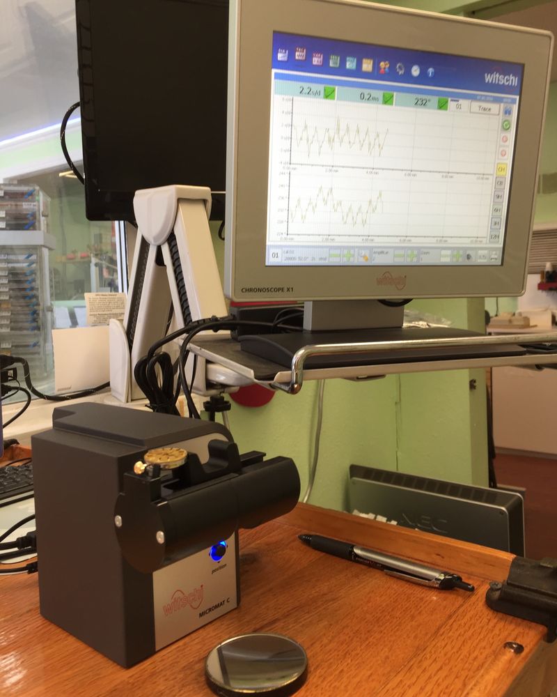 A Witschi Chronoscope X1 timing machine on the workbench, its screen showing rate and amplitude traces, with a watch movement mounted on the Witschi Micromat C microphone stand